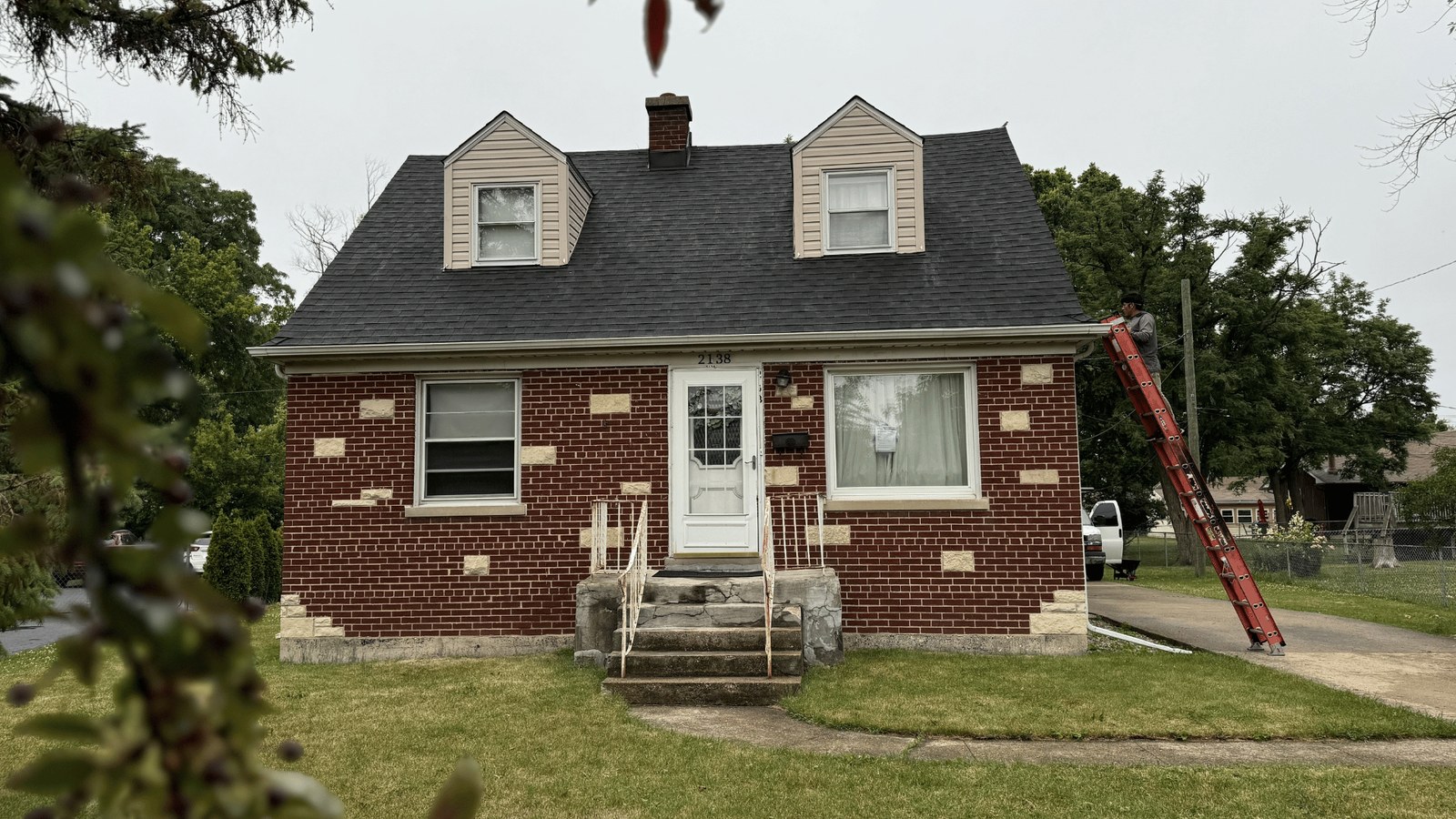 Brick Cape Cod bungalow with a new dark shingle roof, ladder at the eave and the crew truck in the drive