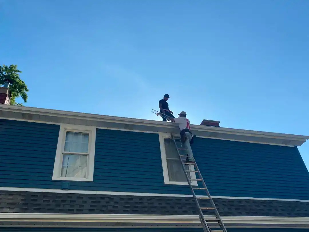 Two crew members on a ladder at the eave of a blue-sided house